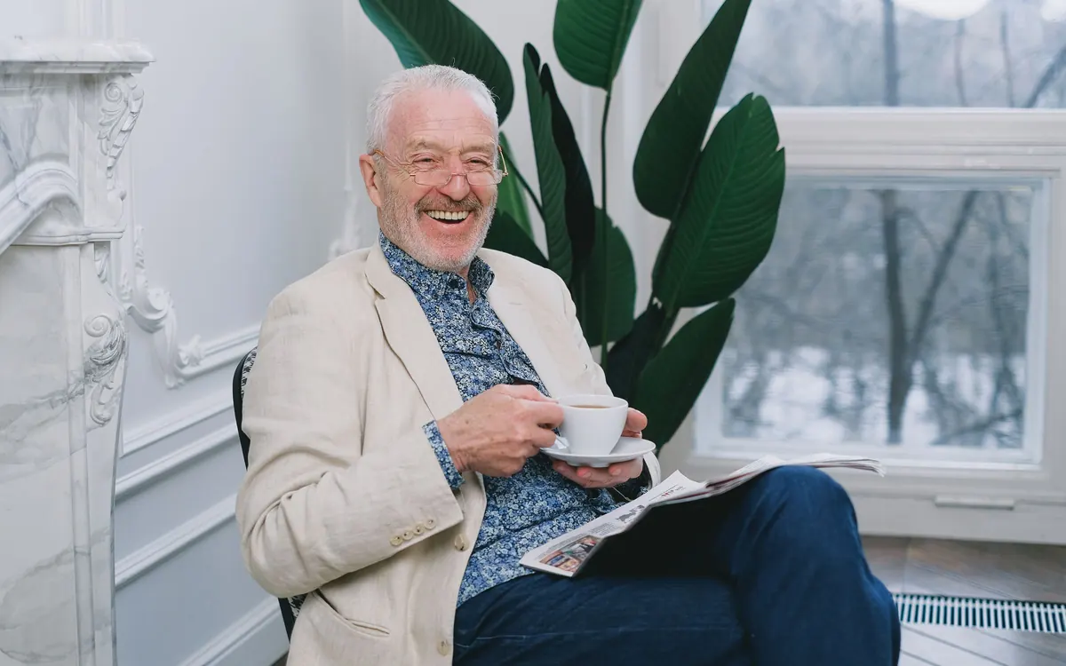 An older man sitting by a bright window with a coffee and a newspaper